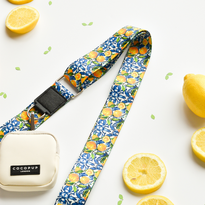 A stylish dog walking accessory, the "Bag Strap - Amalfi Lemon" by Cocopup London features a vibrant yellow strap with a colorful citrus fruit pattern on a white surface. Adorned with slices of yellow lemons, it's paired with a small white pouch featuring a zipper and the "Cocopup London" brand label.
