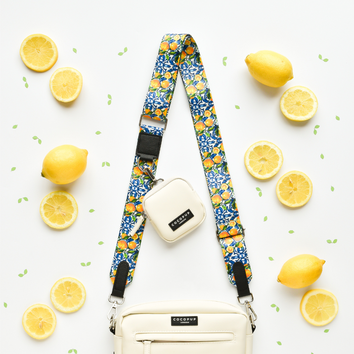 A white handbag featuring the Cocopup London Bag Strap - Amalfi Lemon and a small matching pouch is displayed on a white background. The scene is decorated with whole and sliced lemons, as well as green leaf confetti, creating a fresh and vibrant look.
