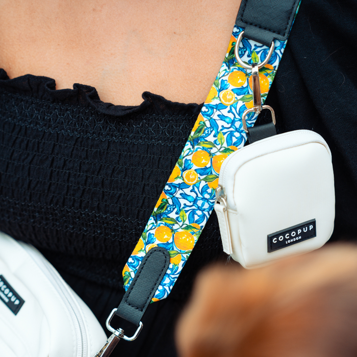 Close-up of a person wearing black clothing with a vibrant yellow strap featuring orange fruit patterns on their shoulder. Attached to the Bag Strap - Amalfi Lemon is a small white bag labeled "Cocopup London." Another white bag, possibly a dog walking accessory, is partially visible on the side.