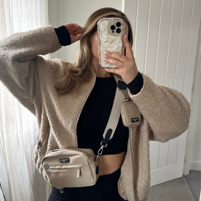 Person in a beige teddy jacket and black crop top holds a tan Cocopup London Dog Walking Bag while taking a mirror selfie.