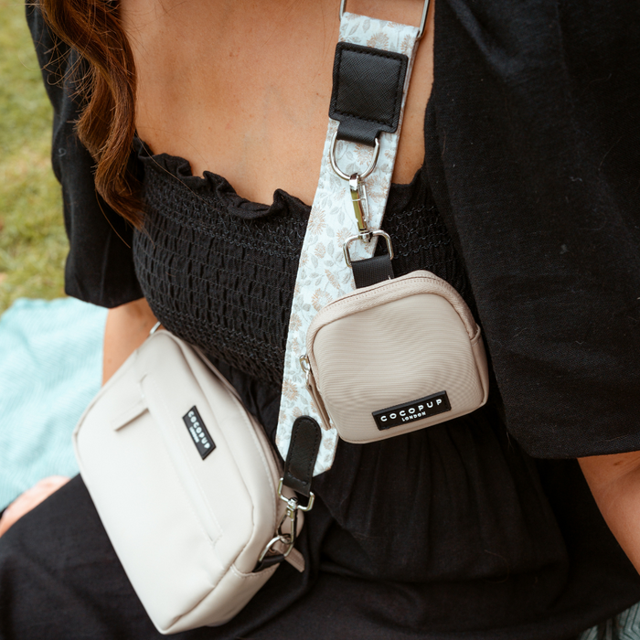 A person wearing a black dress is seated with two crossbody bags. One is beige, worn over the shoulder with a wide patterned strap featuring light-colored designs labeled "Luxe Bag Strap - Garden Meadow" by Cocopup London. The other smaller bag, perfect for dog walks, is attached to the strap and has a label reading "SODOPOP.