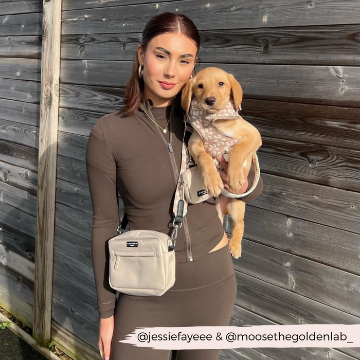 Woman in brown holds puppy in harness, next to mocha flower print Cocopup London dog walking bag by a wooden fence.