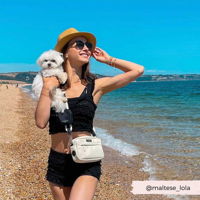 Woman in black lace outfit and hat holds a small white dog and an oyster white Cocopup London dog walking bag on the beach.