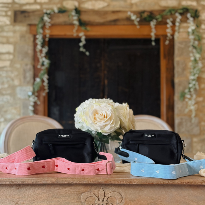 Two black Cocopup London handbags with pink heart-patterned cord straps on a wooden table with white roses.