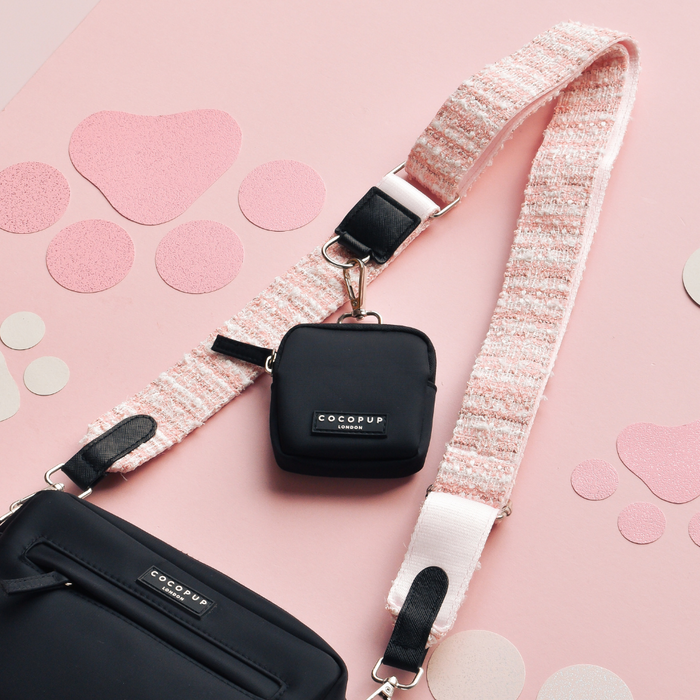Black crossbody dog walking bag with a pink textured tweed strap on a pink background with paw prints.