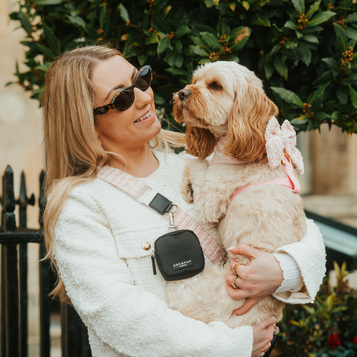 Woman in sunglasses smiles with her fluffy dog, wearing a pink textured tweed Cocopup London bag strap.