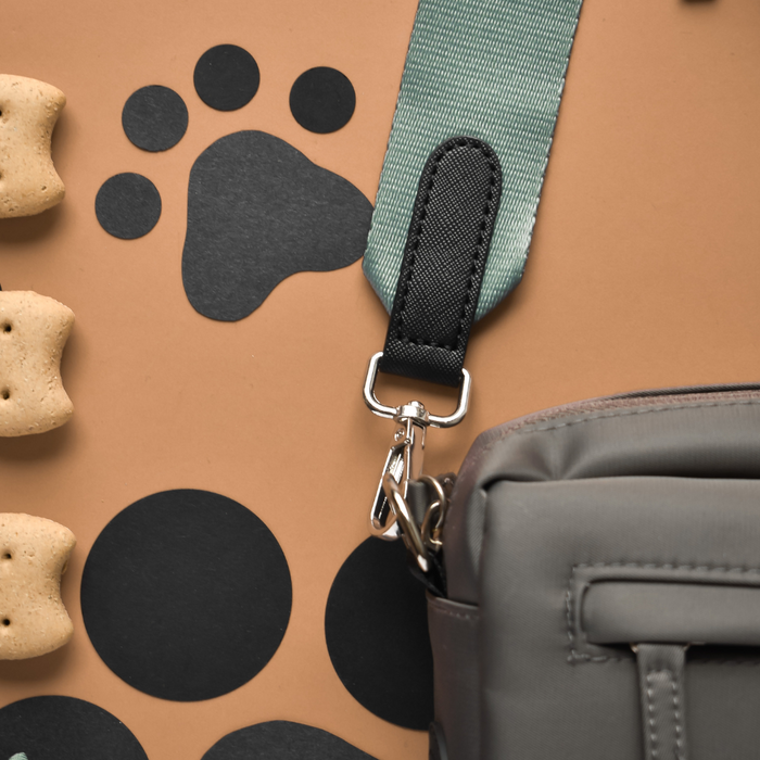 Khaki Cocopup dog walking bag with green strap, placed on tan surface with dog treats and black paw prints nearby.