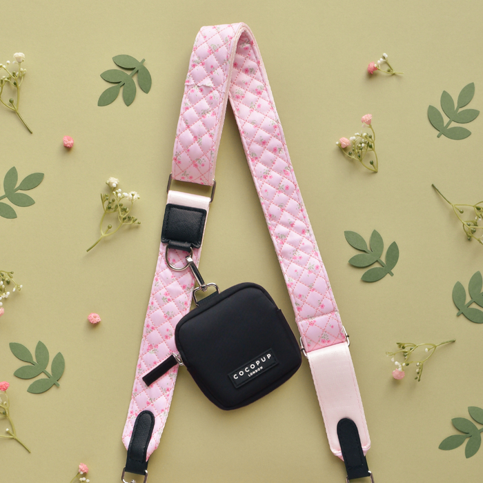 Black pouch with a pink quilted floral Cocopup London strap, placed on beige with flowers and green leaves.
