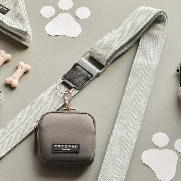 The Dog Walking Mocha Bag Bundle in Sage Tweed by Cocopup London is artfully displayed on a light gray surface, adorned with dog bone-shaped treats and cut-out paw prints. The leash, featuring a sophisticated herringbone pattern and equipped with a sturdy silver clasp, also conveniently doubles as a treat pouch for ease during walks.