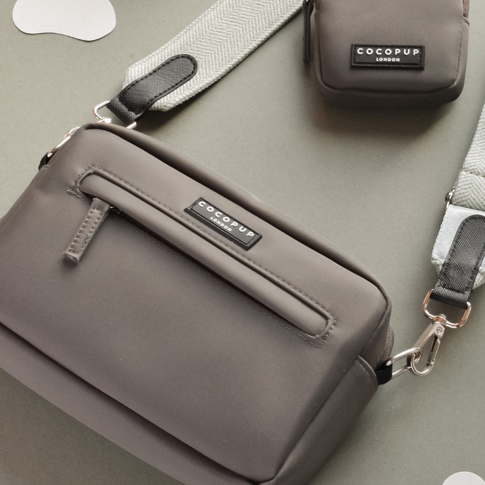 A Dog Walking Mocha Bag Bundle in Sage Tweed, featuring a gray crossbody bag with matching wallet and light gray straps, both branded with "Cocopup London," is placed on a light gray surface. The bag includes a zipper and a small front pocket, and also serves as an elegant dog walking bag. The image highlights the sleek, minimalist design and superior quality of these products.