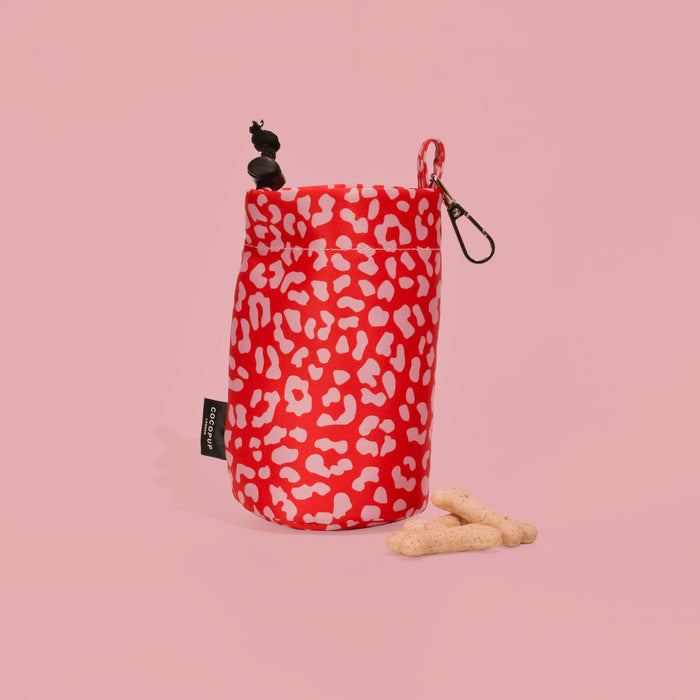 Red and pink leopard print drawstring treat pouch with two bone-shaped treats on a pink background.