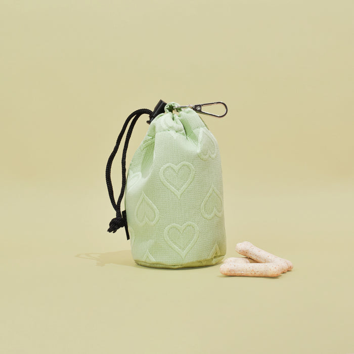 Sage green drawstring treat pouch with heart pattern, next to two bone treats on a beige surface.