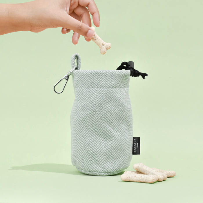 Sage tweed drawstring treat pouch from Cocopup London, filled with treats, with a hand holding a bone-shaped treat above it.