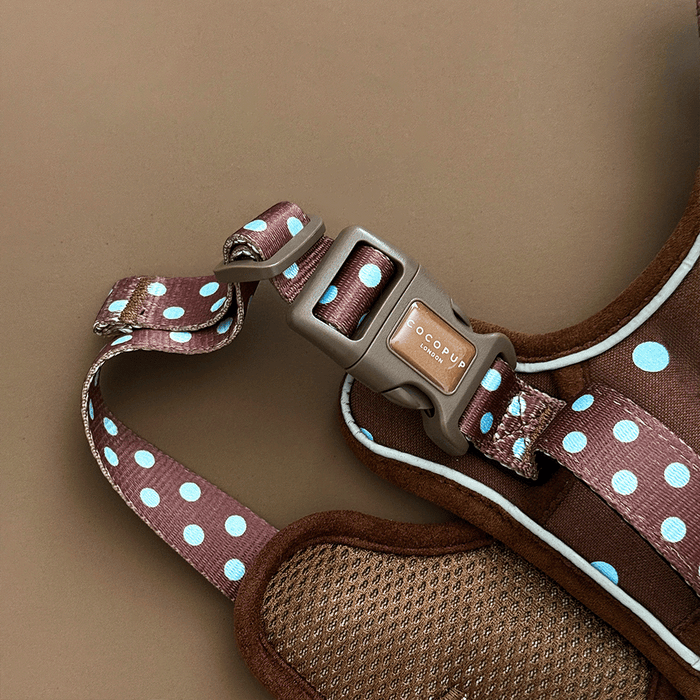 A close up of the buckle on a brown dog harness with light blue polka dots, laid flat on a solid brown background.