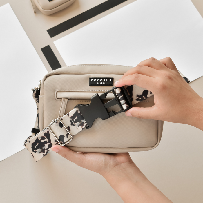 Hands attaching an ivory tortoiseshell bum bag strap to a beige dog walking bag against a neutral background.