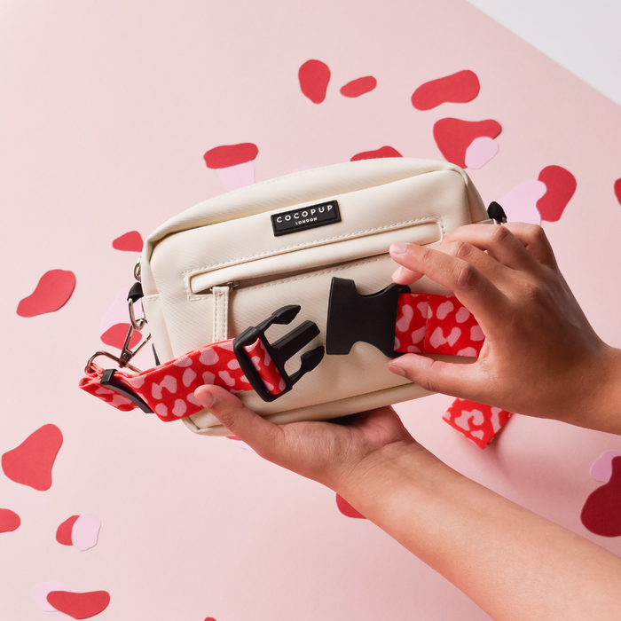 Hands attach a red and pink leopard Cocopup London bum bag strap to a cream crossbody on a pink heart-patterned surface.