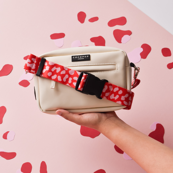 Hand holding a Cocopup London dog bag with a red and pink leopard print strap against a pink background.