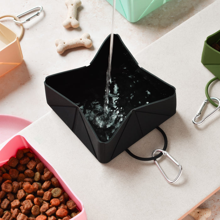 Black Cocopup London foldable travel bowl being filled with water, with dog treats and kibble bowls nearby.
