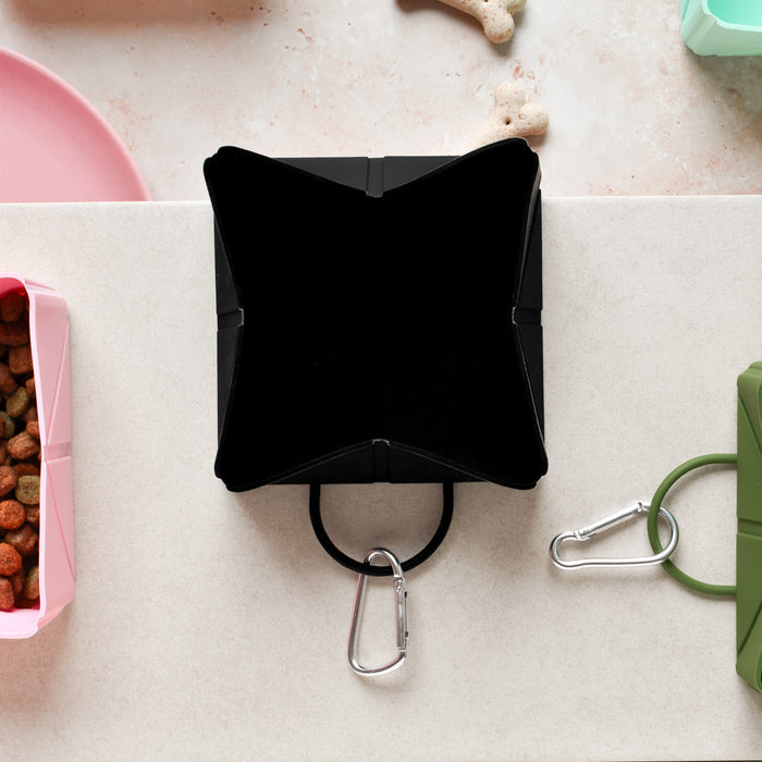 Black Cocopup London foldable travel bowl with carabiner, displayed alongside pet food, treats, and assorted bowls.
