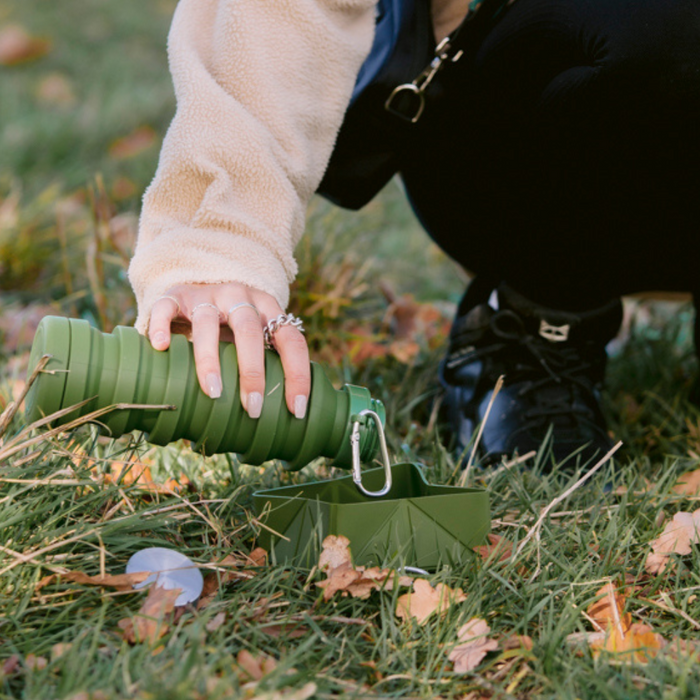 Person pours water from a khaki Cocopup London foldable travel bowl into a matching container on grass.