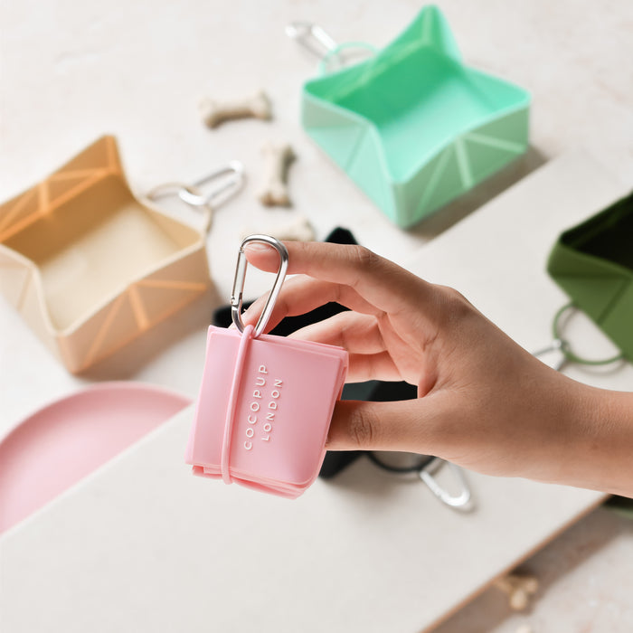 A hand holds a pink foldable travel dog bowl with a carabiner; extra bowls and treats sit in the background.