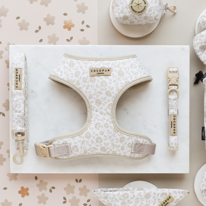 A flat lay image features the Luxe Garden Meadow Adjustable Neck Harness, Lead & Collar Bundle by Cocopup London. The set includes an adjustable neck harness, leash, collar, and waste bag holder. All items are in beige with a subtle floral pattern, displayed on a white marble surface decorated with gold and beige floral motifs.