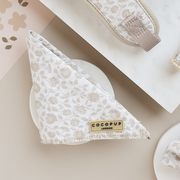 A Luxe Bandana - Garden Meadow in beige and white floral pattern, folded into a triangle, is placed on a white surface. Attached to the bandana is a gold label reading "Cocopup London." Nearby are other items from the Cocopup range with similar patterns and colors, including a Garden Meadow print pouch and part of a card.