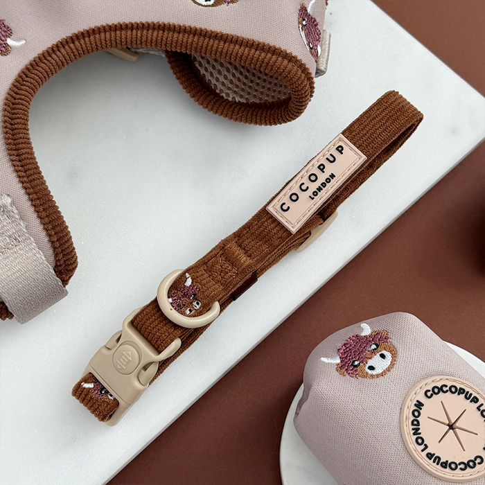Brown and white Highland cow print dog collar, matching harness, and poo bag holder on a marble and brown surface.