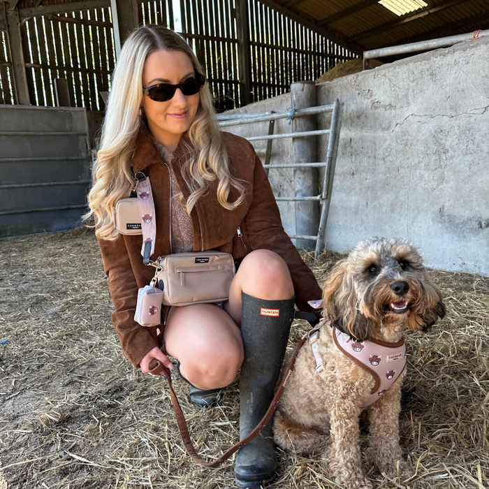 Woman in brown jacket kneels by dog in pink harness with Cocopup London Bag Strap featuring a cow print, inside a barn.