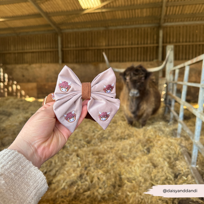 Hand holding a pink Harold the Highland Cow Sailor Bow Tie by Cocopup London, pony blurred in barn background.