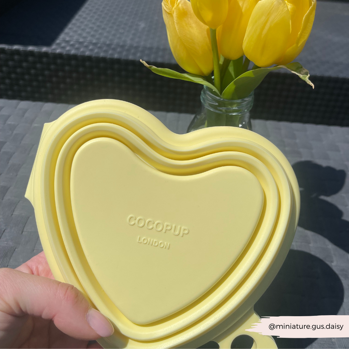 Hand holding three stacked butter yellow collapsible bowls with heart designs beside yellow tulips on a gray table.