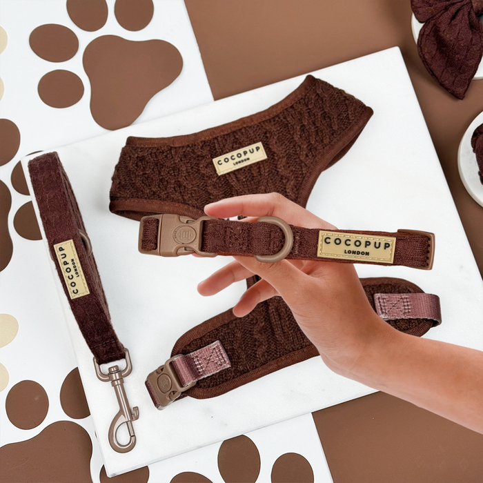 Hand holds brown cable knit harness, lead, and collar set on a white surface with paw prints.