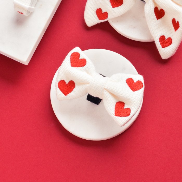 White dog bow tie with red hearts, knit texture, displayed on a red background.