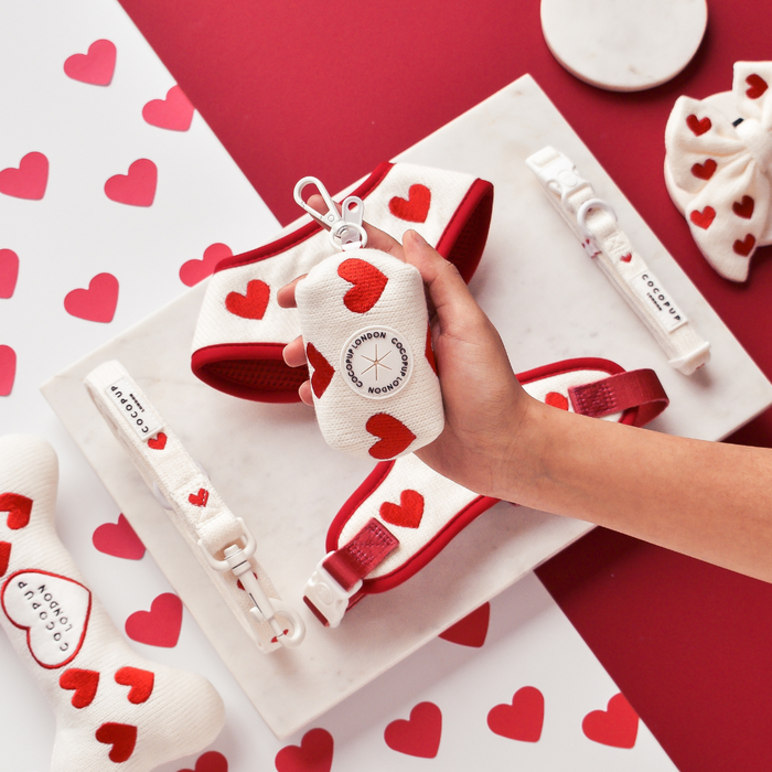 Hand holds a pink knitted Cocopup London poop bag holder; matching accessories rest on a heart-patterned tray.