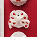 Red and white knitted sailor bow tie with heart pattern, displayed on a round white stand against a red background.