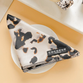 A lightweight and breathable Ivory Tort Bandana from the Cocopup London collection is neatly folded on a white plate placed on a light beige surface. The bandana features a soft texture with black, brown, and white patterns resembling cow spots, marked by the Cocopup London label.