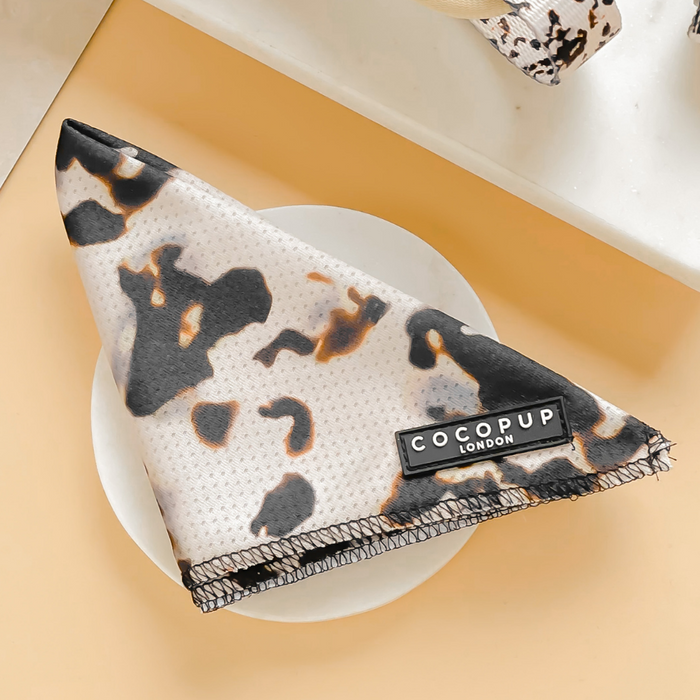 A lightweight and breathable Ivory Tort Bandana from the Cocopup London collection is neatly folded on a white plate placed on a light beige surface. The bandana features a soft texture with black, brown, and white patterns resembling cow spots, marked by the Cocopup London label.