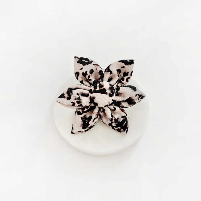 Ivory tortoiseshell-patterned fabric dog collar flower by Cocopup London displayed on a round white surface.