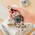 A hand holds a cream and tortoiseshell Cocopup poop bag holder made of soft neoprene; shoes are blurred in the background.