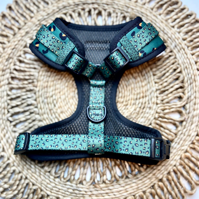 A khaki-colored neoprene dog harness with black padding and a leopard print design lies on a woven, round placemat. The Khaki Leopard Adjustable Neck Harness from Cocopup London includes adjustable straps and black plastic buckles.