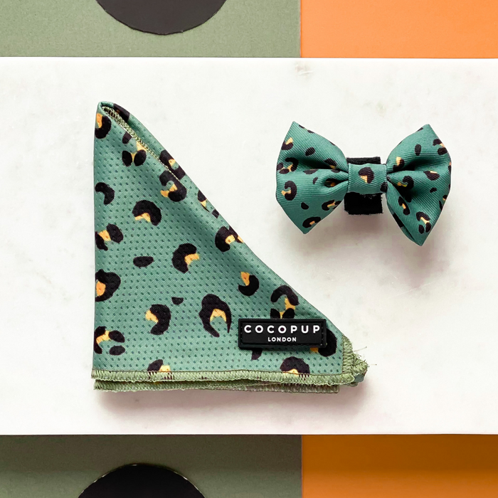 A khaki leopard bandana and matching bow tie with a black and orange cheetah print pattern are displayed on a white and orange background. The bandana, one of the stylish Cocopup London Bandanas, features a black label with white text that reads "COCOPUP LONDON.