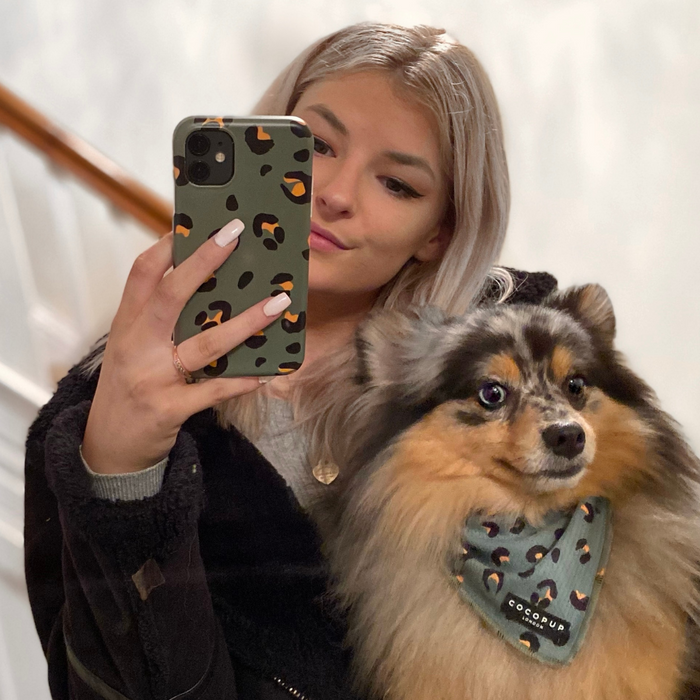 A woman with platinum blonde hair takes a mirror selfie with her phone, which has a green case adorned with leopard spots. She is holding a fluffy tricolor dog wearing a khaki leopard bandana from Cocopup London. The background is blurred, drawing attention to the woman and her dog.