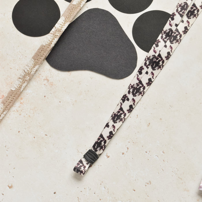 Ivory tortoiseshell Cocopup London lanyard, black paw print paper, and beige textured strip on a speckled surface.
