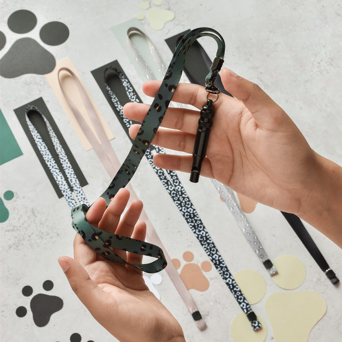 Hands hold a green patterned dog leash with whistle; a khaki leopard print Cocopup London lanyard lies nearby.