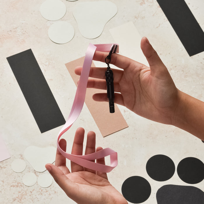 Hands holding a Cocopup London baby pink lanyard with black whistle, against a light background with colored paper accents.