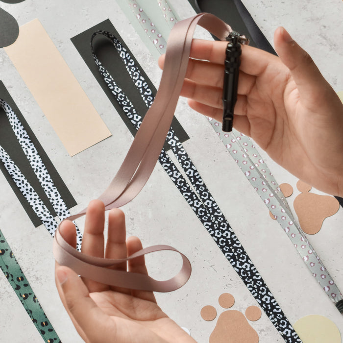 Hands holding a plain nude Cocopup London lanyard, with various patterned lanyards visible in the background.