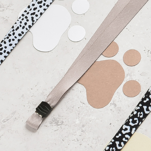 Plain nude lanyard with patterned ribbons and paper cutouts on a lightly textured surface.