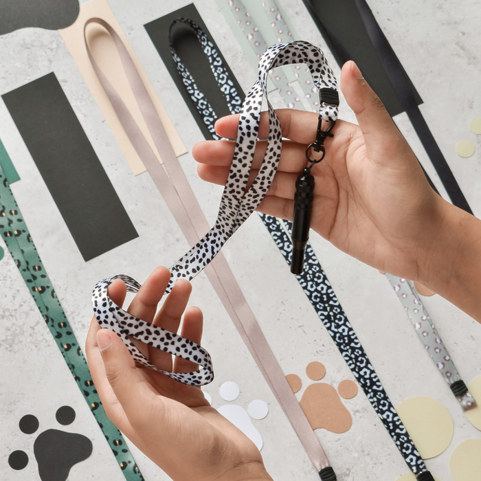 Hands hold a black and beige Cocopup London lanyard with attached silver training whistle over paw print background.