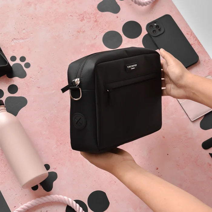 Hands holding a black Cocopup London dog walking bag over a pink desk with a phone and water bottle nearby.