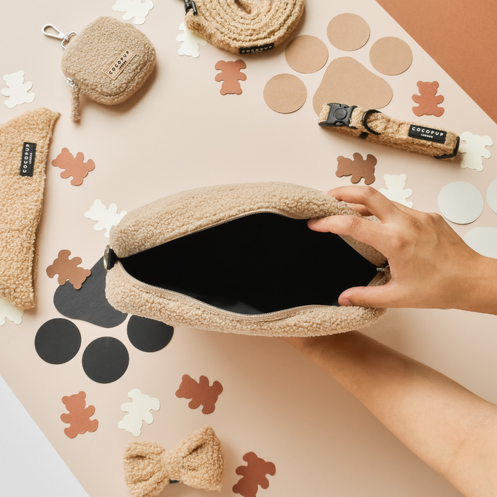 Hand opening a beige Cocopup London Teddy Rupert dog walking bag bundle with matching plush accessories on a table.